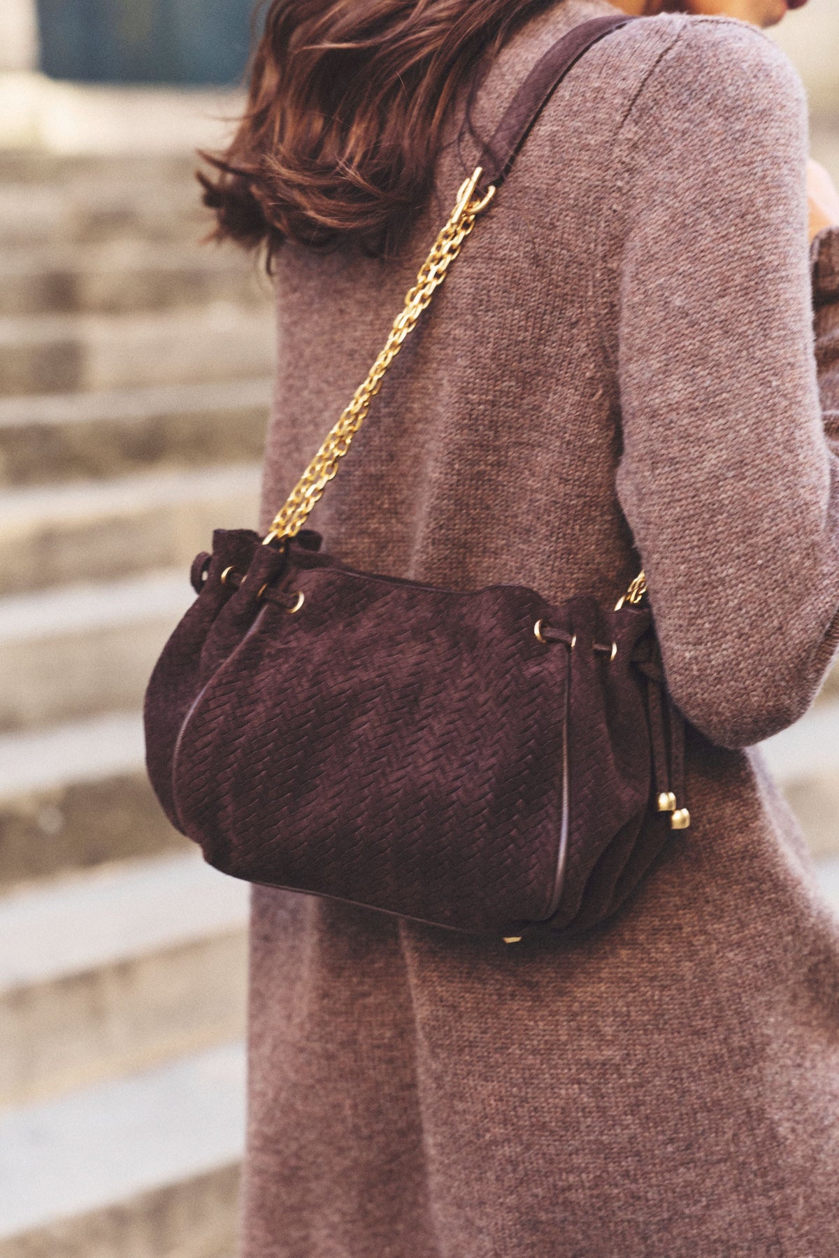 A person wearing a brown outfit stands on steps, carrying a dark brown cuir velours tressé sac bandoulière with a gold chain strap over their shoulder.