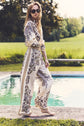 A woman in sunglasses and a patterned white outfit stands barefoot by a pool, with green grass and trees in the background on a sunny day.