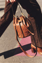 Close-up of a person walking outdoors, holding a brown sac seau cuir with gold hardware. The sac bandoulière femme is styled elegantly against dark brown pants and heeled shoes, illuminated by natural sunlight.