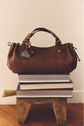 A brown sac à main in cuir d’agneau tressé with gold hardware sits on a stack of large books atop a wooden stool. Showcasing the savoir-faire de la Maison, the bag has a leather tag and is arranged indoors on a wooden floor against a neutral wall.