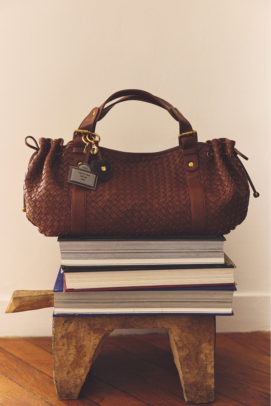 A brown sac à main in cuir d’agneau tressé with gold hardware sits on a stack of large books atop a wooden stool. Showcasing the savoir-faire de la Maison, the bag has a leather tag and is arranged indoors on a wooden floor against a neutral wall.