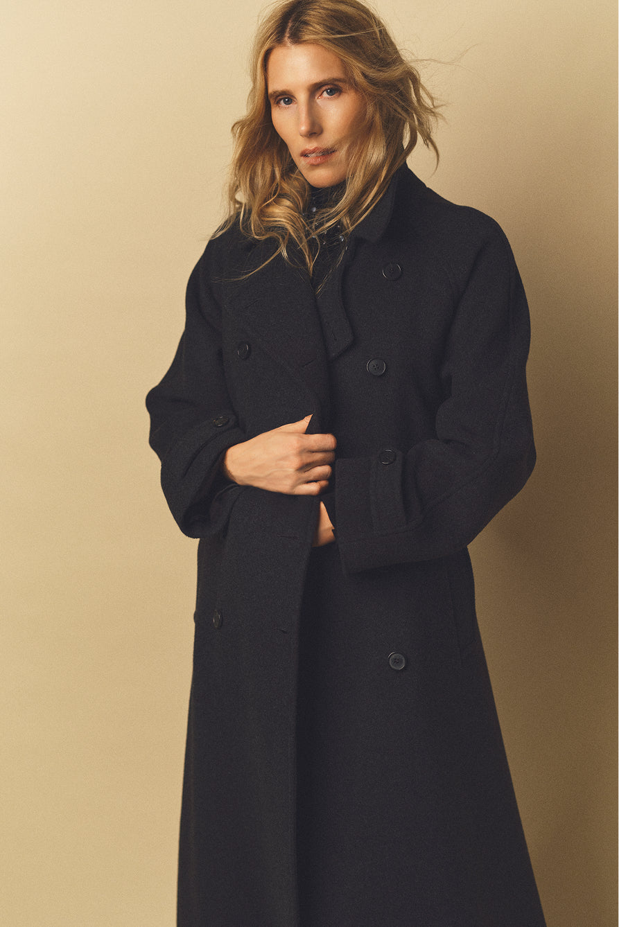A woman with wavy blonde hair wears a chic manteau long femme, a black double-breasted coat reminiscent of a classic trench laine, and looks at the camera against a plain beige background.