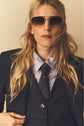 A person with long blonde hair wears a veste rayée dark suit, polka dot tie, striped shirt, and aviator sunglasses, looking confidently at the camera against a beige background.