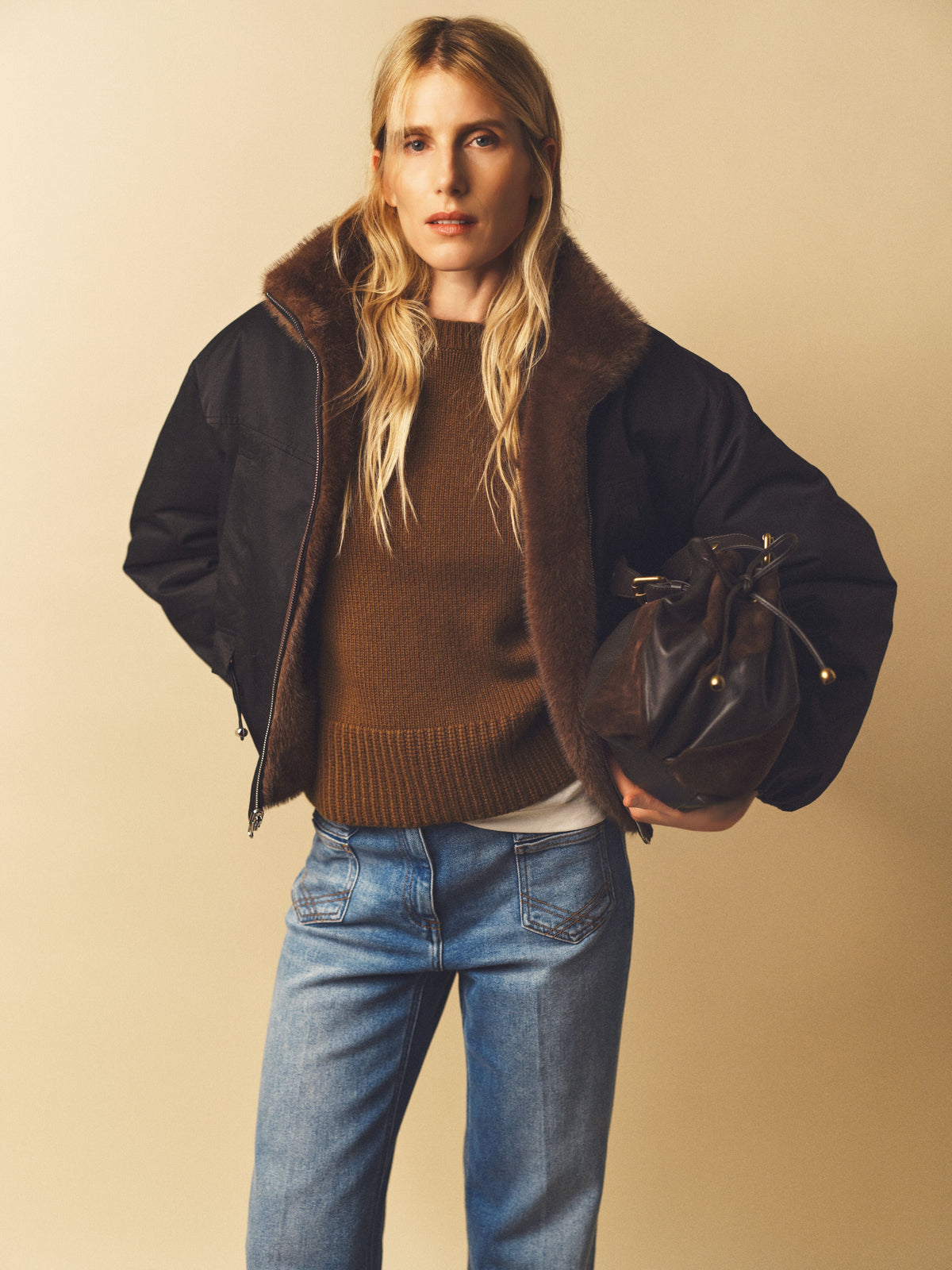 A woman with long blonde hair wears a brown sweater, blue jeans, and a black parka courte with a fausse fourrure collar, holding a dark brown drawstring bag against a beige background.