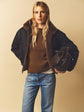 A woman with long blonde hair wears a brown sweater, blue jeans, and a black parka courte with a fausse fourrure collar, holding a dark brown drawstring bag against a beige background.