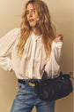 A woman in a white chemise en crêpe with col volanté and manches bouffantes poses in blue jeans, holding a black woven handbag, her long wavy blonde hair falling gracefully against a neutral background.