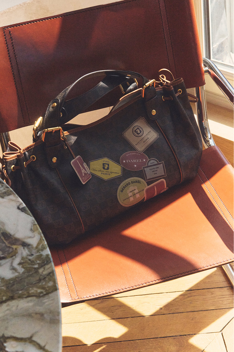 A dark sac Le 24H designer handbag decorated with travel-themed patches and tags sits on a brown leather chair in sunlight near a marble table, showcasing the timeless allure of maroquinerie Darel.