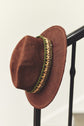 A brown chapeau de soleil made of paille tressée with a decorative green and gold band rests on a black railing against a light background.