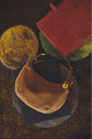 Brown suede handbag in cuir velours with a gold clasp rests on a black book atop a round table with a textured yellow-green surface; a leather folder sits nearby on another table, evoking an air of chic parisienne elegance.