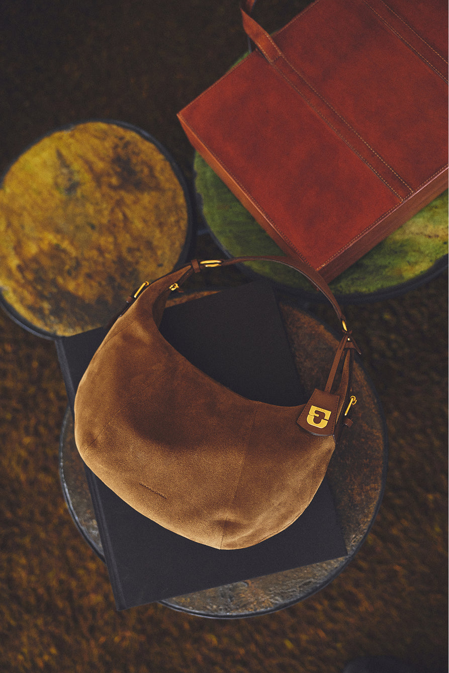 Brown suede handbag in cuir velours with a gold clasp rests on a black book atop a round table with a textured yellow-green surface; a leather folder sits nearby on another table, evoking an air of chic parisienne elegance.