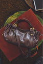 A brown cuir de vachette sac à main with gold hardware rests atop a closed, red leather-bound book, both placed on a green circular surface.