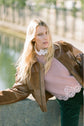 A woman with long blonde hair, wearing a brown jacket and a pink pull dentelle sweater, leans on a concrete ledge near a reflective body of water outdoors.