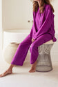A person in a bright pyjama violet deux pièces sits barefoot on a wire stool next to a large beige vase, with a white bathtub in the background. Their face is not visible.