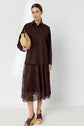 Woman wearing a brown chemise en popeline, a brown lace midi skirt, and brown heeled sandals. She holds a woven tan handbag and stands against a plain white background.