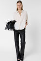 A woman wearing a loose white blouse and black leather pants holds a black fringe bag. She stands facing forward against a plain white background.