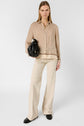 A woman stands facing forward, wearing a patterned beige blouse, cream wide-leg pants, and black shoes. She holds a black handbag in her left hand against a plain white background.