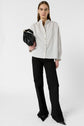 A woman stands facing forward wearing a white chemise Calixte à col mao, black pants, and pointed black shoes, holding a black handbag under her arm. The background is plain white.