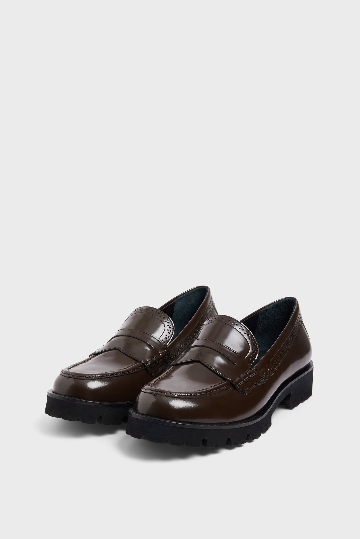 A pair of shiny dark brown mocassins cuir with a semelle crantée in black chunky rubber, displayed on a light gray background for a style classique.