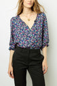 A woman wearing a blue floral blouse with a V-neck and black pants, accessorized with layered gold necklaces, stands against a plain light background.