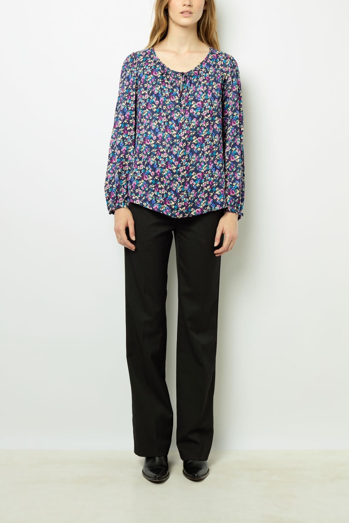 A person stands against a white background wearing a long-sleeve, floral-patterned blouse, black pants, and black shoes.