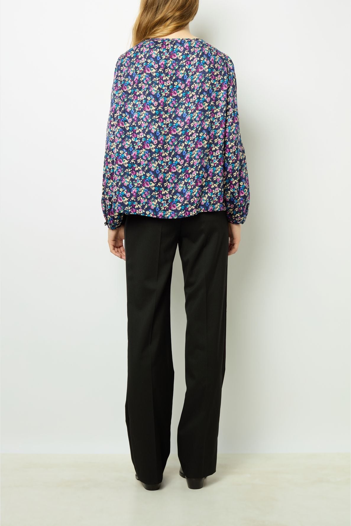 A person with long hair stands facing away, wearing a dark floral blouse with long sleeves and loose-fitting black pants against a plain white background.