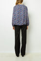 A person with long hair stands facing away, wearing a dark floral blouse with long sleeves and loose-fitting black pants against a plain white background.