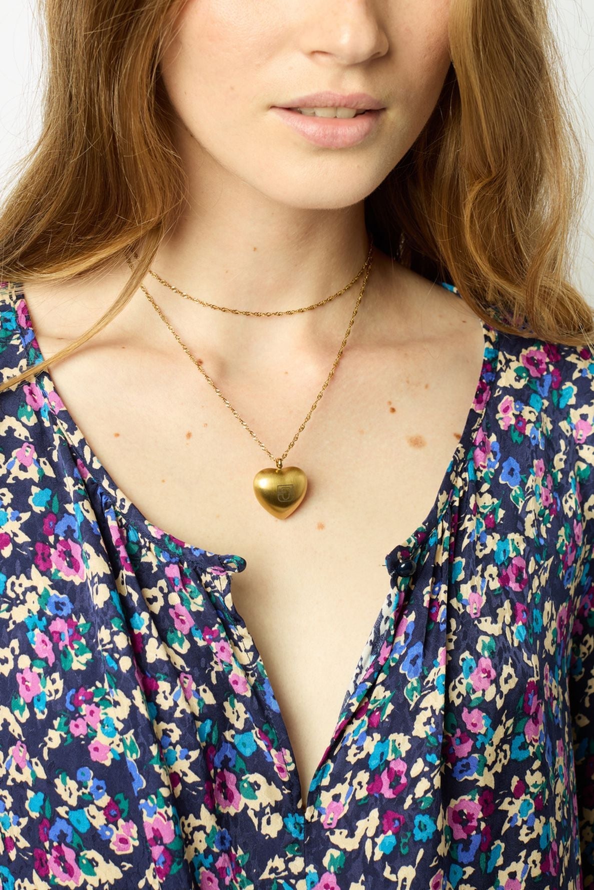 A woman wearing a floral-patterned blouse, layered gold necklaces, and a gold heart locket. Only the lower half of her face and upper torso are shown.