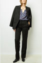 Woman wearing a black suit with a floral blouse underneath, black dress shoes, and a gold necklace. The suit jacket is draped over her shoulders, and she stands with one hand in her pocket against a plain background.