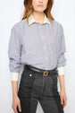A person wears a blue-and-white striped popeline coton chemise rayée with a crisp col blanc and white cuffs, tucked into high-waisted black denim jeans with a black belt. The image is cropped at the shoulders and thighs.