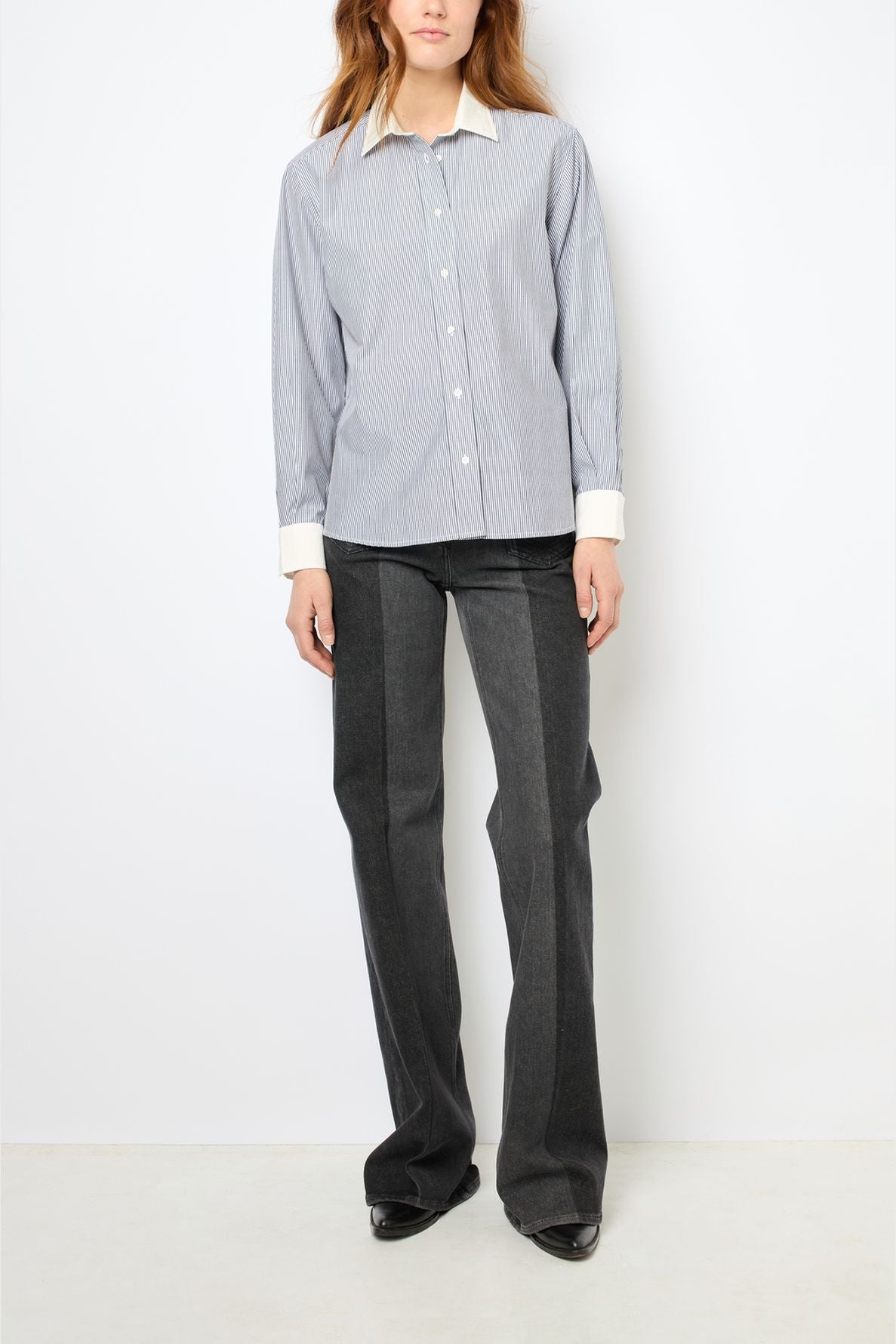 A person stands against a white wall wearing a light blue chemise rayée in popeline coton, featuring a crisp col blanc and cuffs, dark gray flared pants, and black shoes. Their face is partially visible, and their hands rest at their sides.