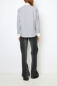 A person stands facing a white wall, wearing a light grey chemise rayée in crisp popeline coton with elegant col blanc and white cuffs, paired with dark grey flared pants. The person’s hands rest at their sides, and only their back is visible.