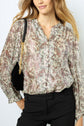 Woman wearing a long-sleeve, floral-patterned blouse and black pants, carrying a black shoulder bag with a gold chain strap, standing against a light background.