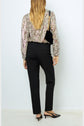 A person stands facing away, wearing a floral long-sleeve blouse, black pants, black slingback heels, and carrying a black shoulder bag with a gold chain strap.