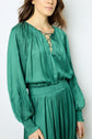 Woman wearing a blouse satin plissé in a long-sleeved, pleated, teal satin dress with a blouse encolure lacée and a smocked waistband, standing against a plain white background. Her face is partially visible.