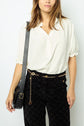 Woman with light brown hair wears a white blouse, black patterned pants, a gold chain belt, and a black shoulder bag, standing against a plain background. Her face is partially visible.