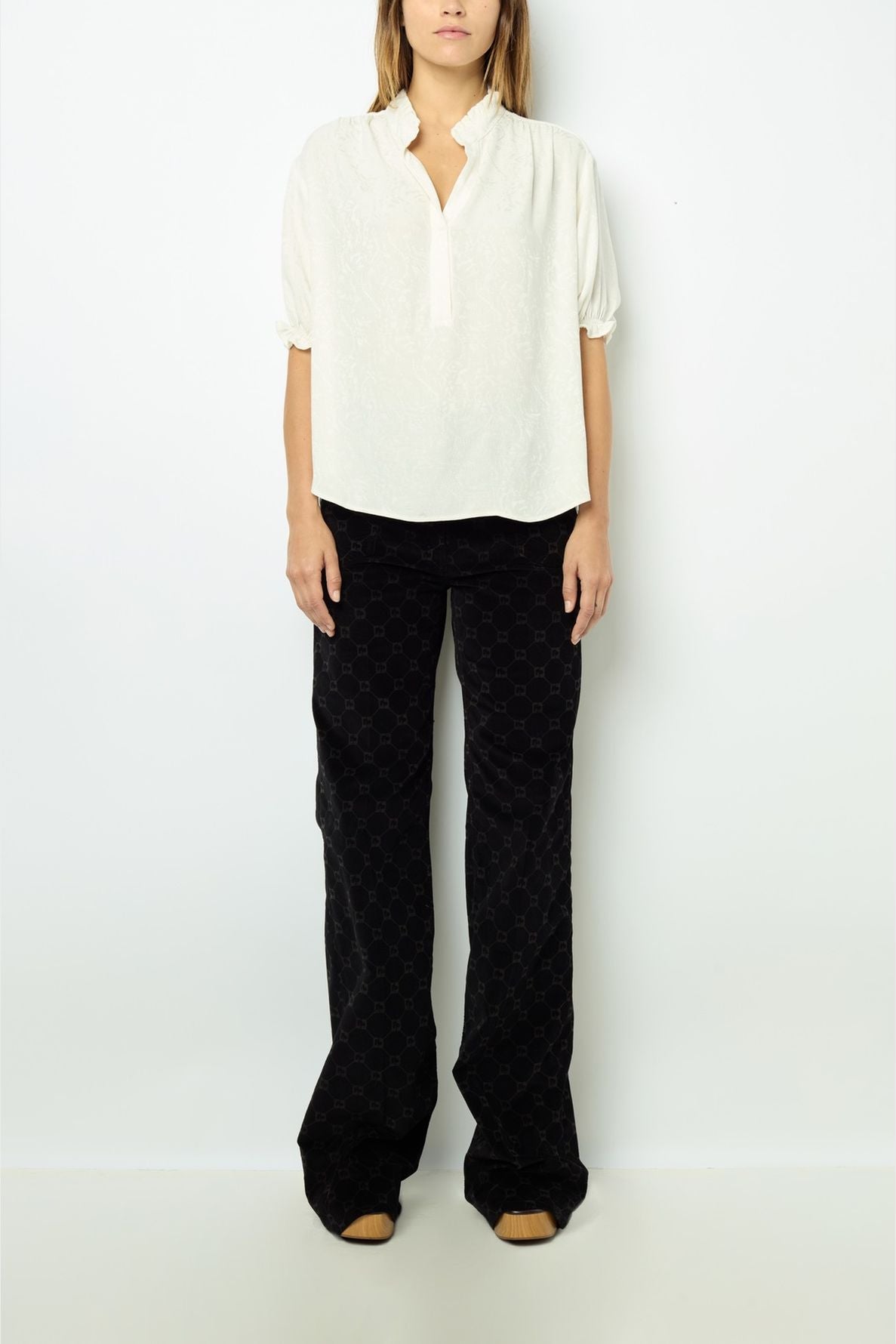 A person stands against a plain white wall wearing a loose white blouse with short sleeves and black wide-leg pants. Their hands are by their sides and their face is not visible.