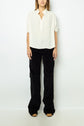 A person stands against a plain white wall wearing a loose white blouse with short sleeves and black wide-leg pants. Their hands are by their sides and their face is not visible.