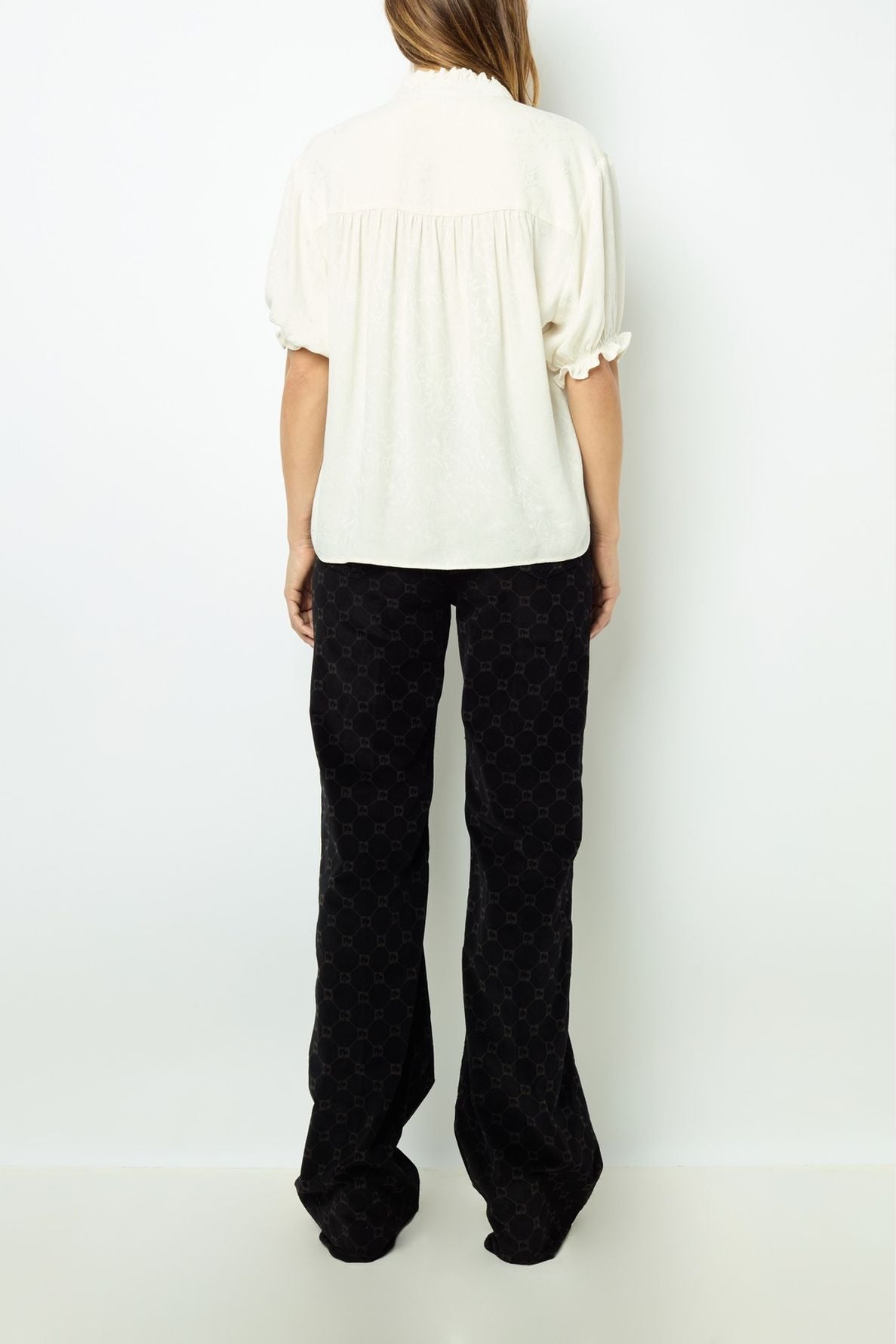 A person stands facing away, wearing a white short-sleeve blouse and loose black patterned pants. The background is plain white.