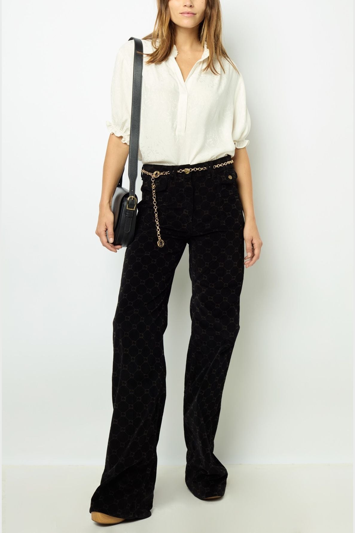 Woman wearing a white blouse, black patterned pants with a gold chain belt, tan shoes, and a black crossbody bag, standing against a plain white background.