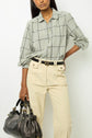 A person wearing a light green plaid shirt and beige high-waisted pants holds a large black leather handbag.