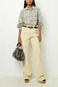 A person wearing a gray plaid shirt, high-waisted beige pants, and black belt, holding a large black handbag, stands against a plain white background.