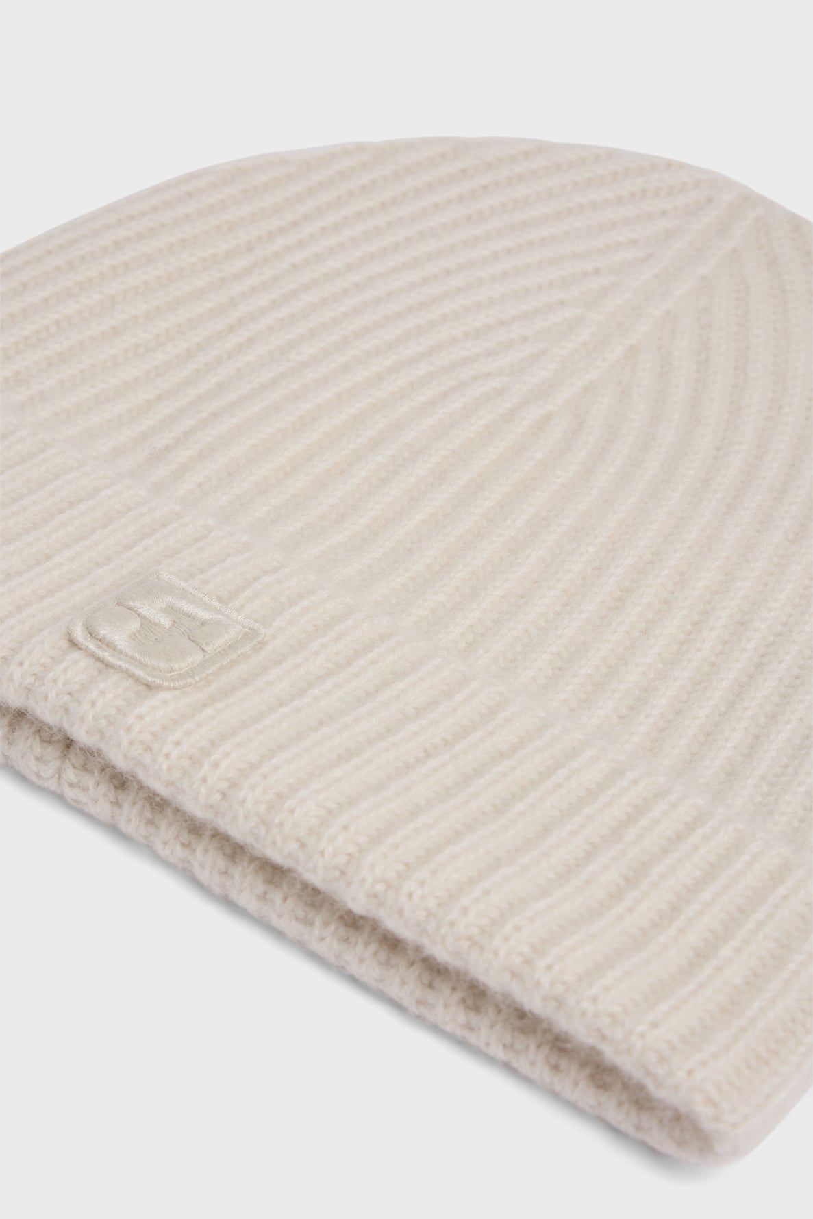 A close-up of a beige ribbed knit beanie hat, or bonnet à côtes, crafted in soft cachemire with a subtle monogramme GD stitched on the cuff, photographed against a white background.