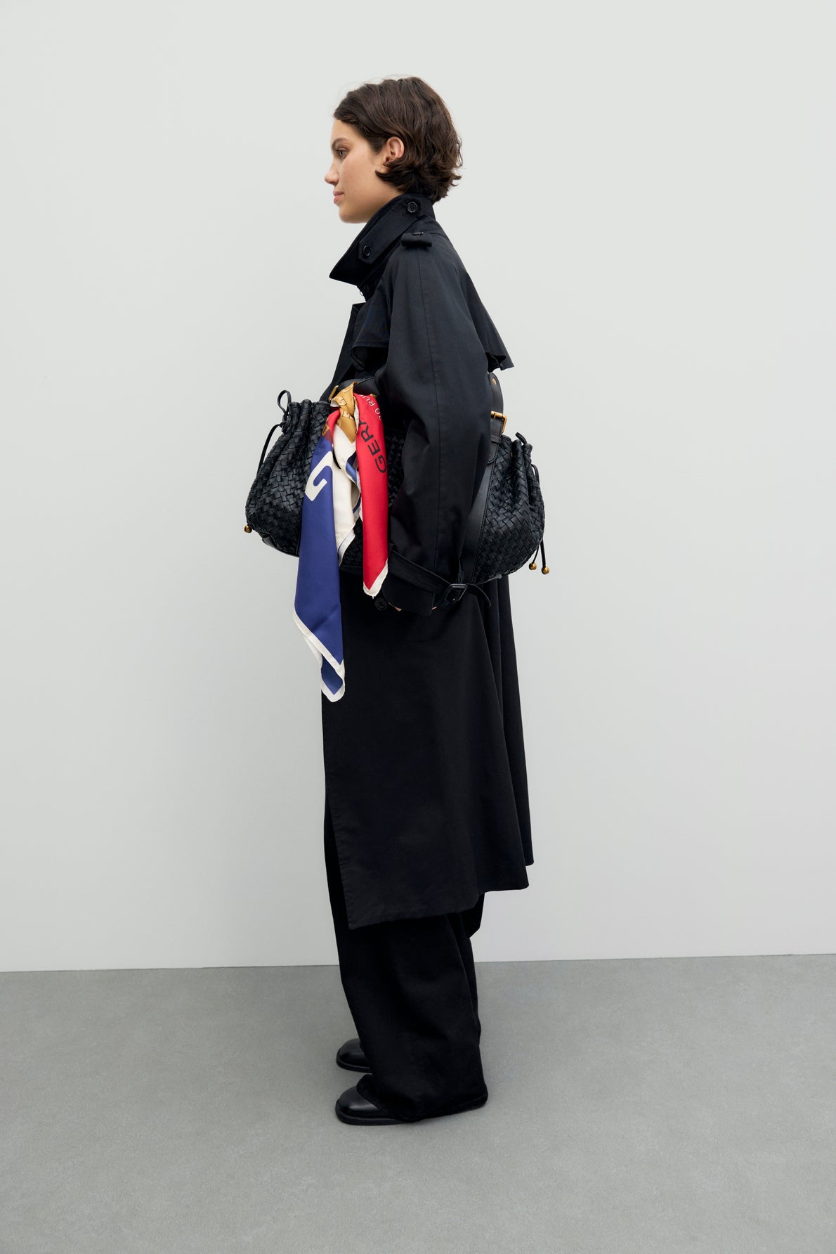 Person in a trench long black coat and wide-leg pants stands sideways, holding a black woven bag with a colorful scarf tied to it. The background is plain and light-colored.