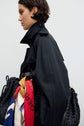 A person in a black trench long manteau femme with short, dark hair carries a woven black bag adorned with colorful scarves. The background is plain and light-colored.