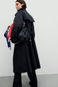 A person in a manteau femme trench long holds a large black woven bag with gold hardware and a colorful scarf tied to it, standing against a plain light-colored wall.