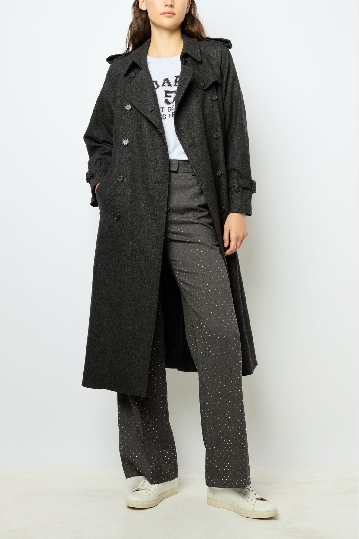 A person wearing a trench long dark coat, gray polka dot trousers, a white graphic T-shirt, and white sneakers stands against a plain light background.