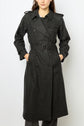 A woman wearing a long, dark gray trench coat with a belt and button details—reminiscent of an élégant trench long or manteau laine femme—stands with her hands in the coat’s pockets against a plain background.