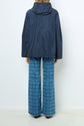Person facing away, wearing a navy parka courte with a capuche and blue wide-leg pants featuring a checkered pattern, crafted using a refined polyester technique, standing against a plain white background.
