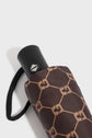 A close-up of a folded brown parapluie pliant with a geometric tan pattern, partially inside its matching sleeve. The handle has a black strap and a silver triangular logo on a circular black button, while subtle monogramme GD details enhance its sophisticated design.