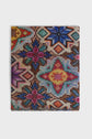 A colorful, square woven textile with intricate geometric and floral patterns in blue, purple, yellow, red, and green on a light background. Reminiscent of a foulard imprimés géométriques, the bottom edge is slightly frayed.